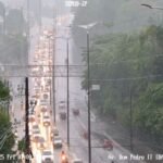 Rua alagada em João Pessoa durante chuva intensa com sinal da Defesa Civil em destaque.