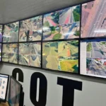 Pessoas em uma sala de monitoramento, observando múltiplas telas que exibem imagens de câmeras de segurança, em um contexto de vigilância eleitoral.