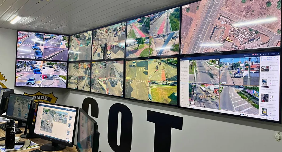 Pessoas em uma sala de monitoramento, observando múltiplas telas que exibem imagens de câmeras de segurança, em um contexto de vigilância eleitoral.