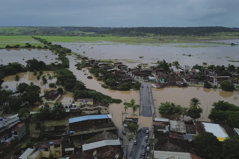 Rua alagada com casas submersas devido a fortes chuvas em Pernambuco ou Paraíba, com equipes de resgate em ação.