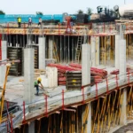 Trabalhadores em um canteiro de obras em João Pessoa, com edifícios modernos ao fundo.