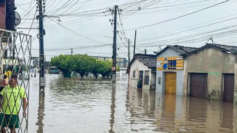 Rua inundada na Paraíba após chuvas intensas, mostrando casas submersas e devastação.