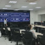 Tela de monitoramento de centro de comando e controle exibindo imagens de câmeras de segurança e dados em tempo real, com equipes de segurança no fundo.