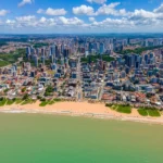 Vista panorâmica aérea da orla de João Pessoa ao amanhecer, destacando praias, mar e a cidade em desenvolvimento.