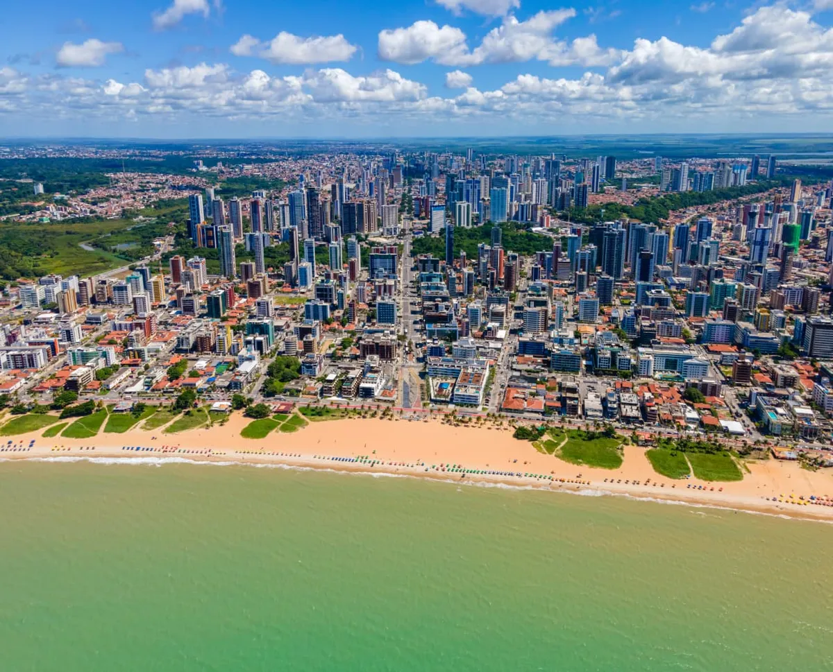 Vista panorâmica aérea da orla de João Pessoa ao amanhecer, destacando praias, mar e a cidade em desenvolvimento.