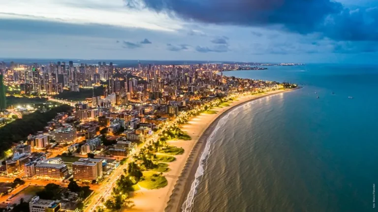 Pôr do sol na Praia de Tambaú em João Pessoa, Paraíba, com coqueiros e edifícios ao fundo.